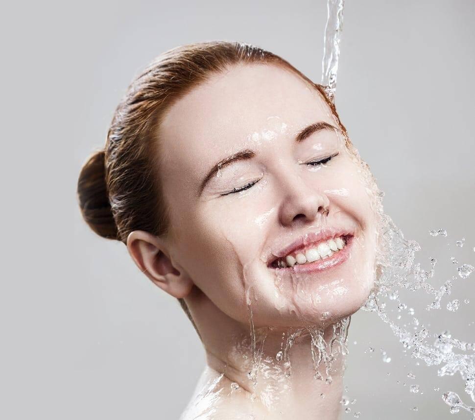 A smiling person with closed eyes enjoys water splashing onto their face, refreshing and cleansing their skin. Their hair is tied back tightly in a bun, and they appear relaxed and content. The background is a soft grey gradient, emphasizing the serenity of the moment.