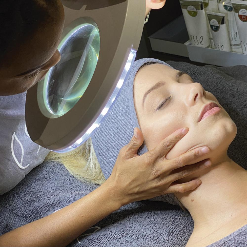 A person receiving a facial examination under a magnifying lamp. The esthetician’s hands are gently holding the client’s face, which is covered partially with a headband. Various skincare products are visible in the background.