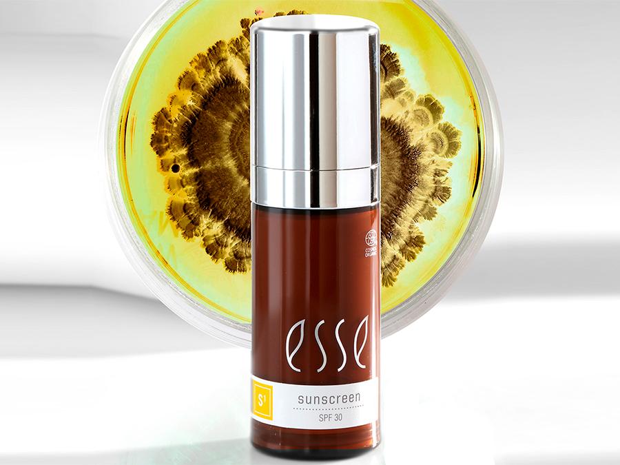 A bottle of Esse sunscreen with SPF 30 is displayed in front of a petri dish containing a yellow and brown pattern. The bottle is brown with a silver pump and white label.