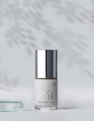 The Esse Eye Contour Cream, featuring a shiny silver cap, stands on a white surface. Soft leaf shadows fall across the light gray background, giving the scene a calm and elegant vibe.