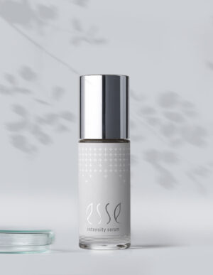 A bottle of Esse Intensity Serum with a shiny silver cap is shown against a light gray background with soft leaf shadows. The minimalist white bottle features small silver crosses and the Esse Intensity Serum name.