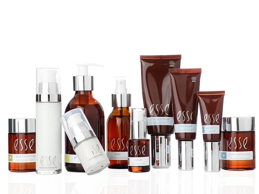 A display of various Esse skincare products in bottles and tubes of different sizes. The packaging features a combination of brown, white, and clear materials with metallic-colored caps, and the brand name "Esse" is prominently displayed on each item.