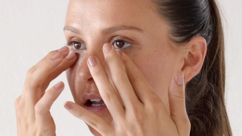 A woman with brown hair pulled back is gently applying Esse Eye & Lip Cream around her eyes, possibly as part of her skincare routine. She has a neutral expression on her face.
