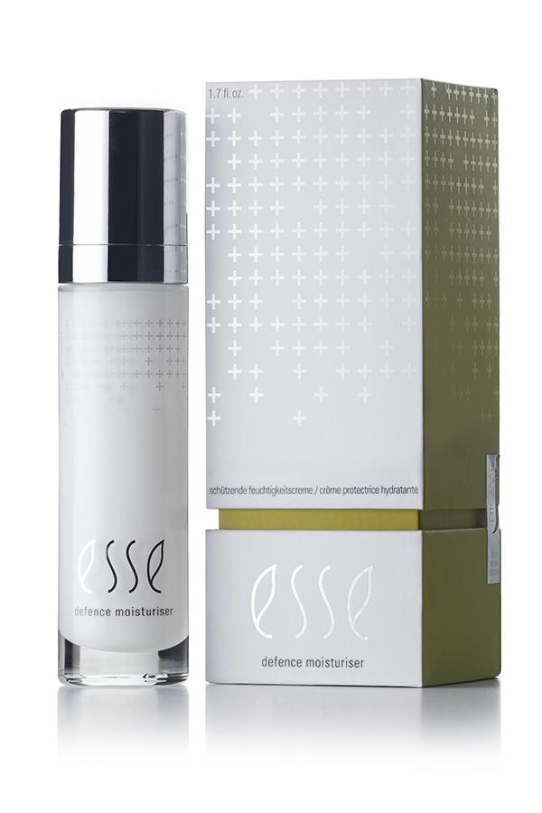 A sleek white bottle of Esse Defence Moisturiser, adorned with a shiny silver cap, stands next to its box. Both the bottle and the rectangular box display the brand's name "Esse" in elegant cursive, complemented by a pattern of small crosses. The box features an elegant color scheme of white and olive green.