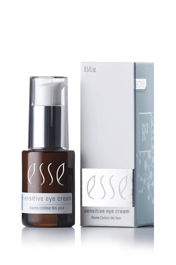 A brown bottle with a silver pump dispenser of "Esse Sensitive Eye Cream" is set in front of its grey and white packaging box. Both the bottle and the box feature the product name along with multilingual text. The bottle has a capacity of 0.5 fl. oz. (15 ml).