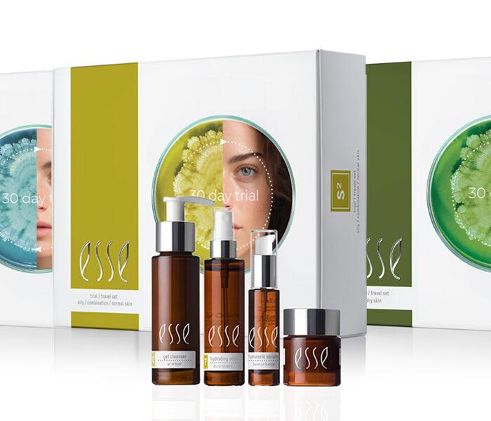 Skincare trial sets in white boxes with green accents, each featuring a partial photo of a woman's face. In front, five brown skincare product bottles and jars labeled "esse" are arranged in a row.