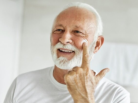 A smiling elderly man with white hair and a white beard is applying face cream to his cheek using his finger. He is wearing a white t-shirt and is standing in a well-lit room with a light background.