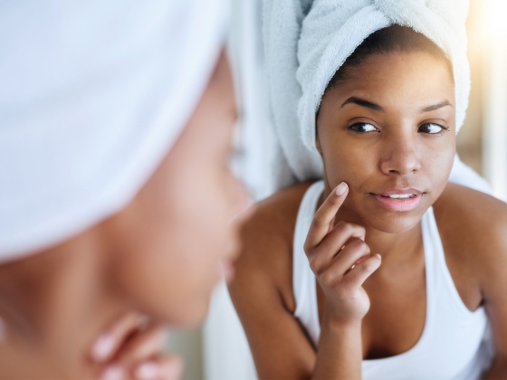 A woman with a white towel wrapped around her head looks at her reflection in a mirror. She is gently touching her cheek with her finger and is wearing a white tank top. The background is softly blurred.
