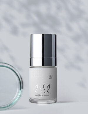 A bottle of Import placeholder for 58570 probiotic serum with a metallic silver cap stands upright against a light background, creating soft shadows. A glass lid is partially visible on the left side.