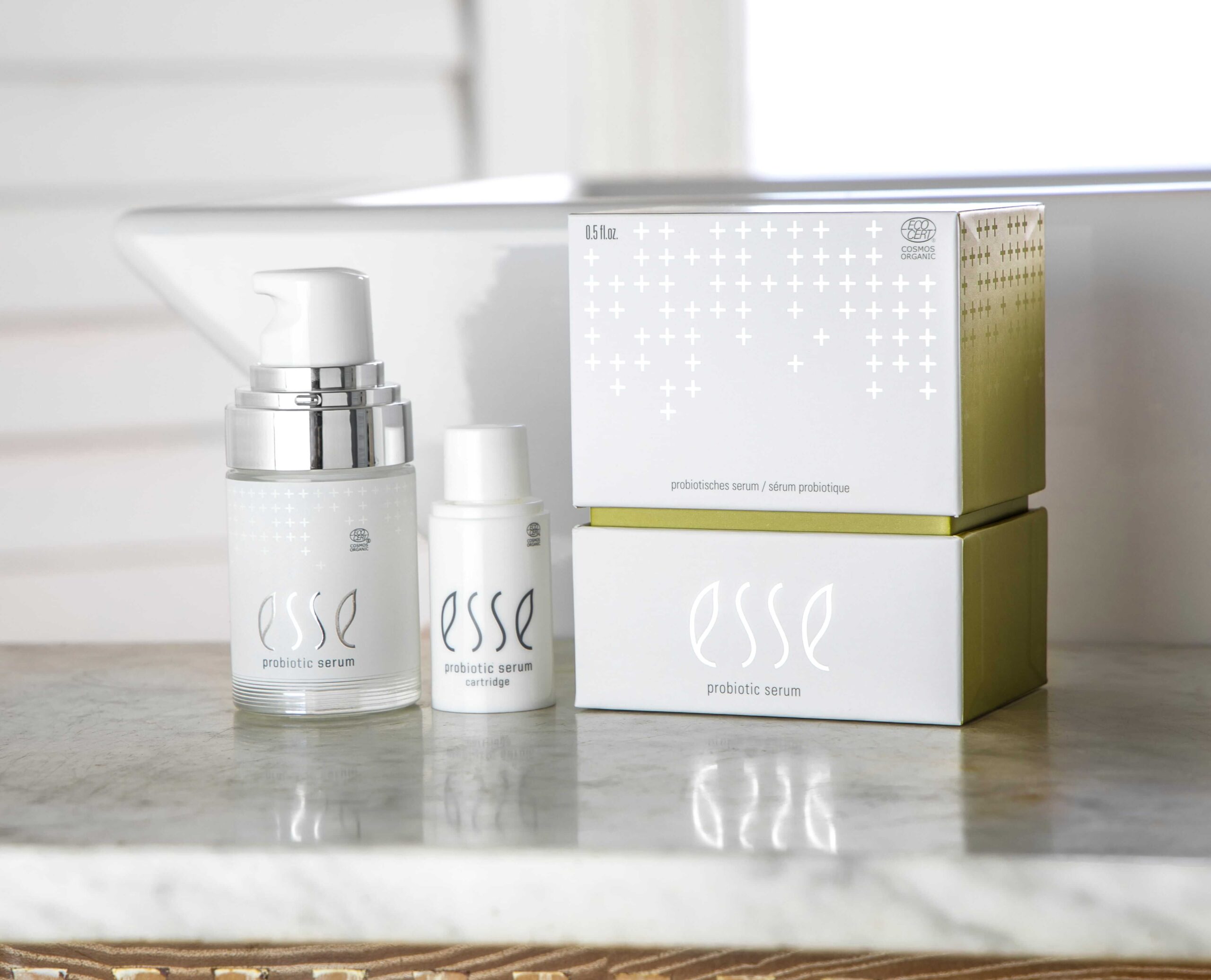 Two bottles and two boxes of Esse Probiotic Serum, featuring white and metallic minimalist packaging, are arranged on a marble worktop near a window.